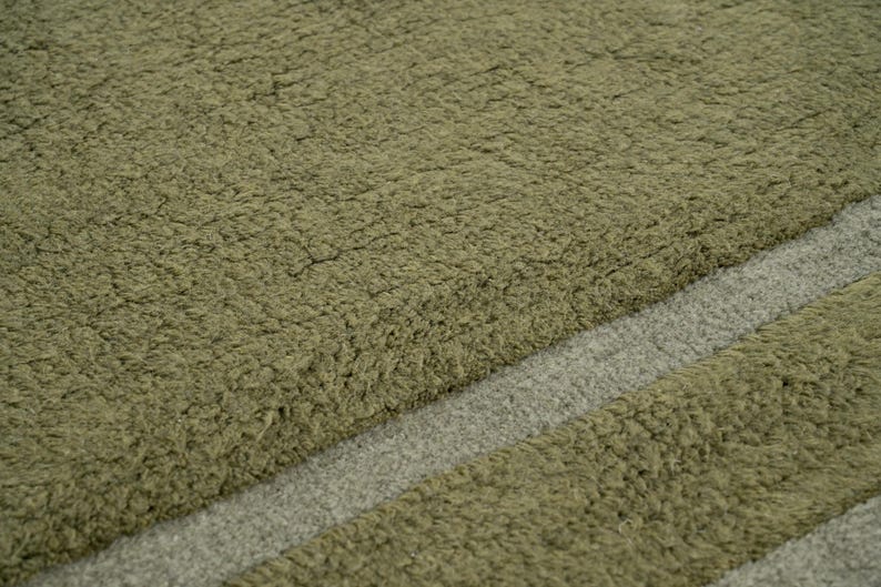 Pode incluir: Close-up de um tapete verde-oliva texturizado com uma faixa diagonal cinza claro. O tapete tem uma apar&ecirc;ncia macia e felpuda, sugerindo uma textura macia e confort&aacute;vel. O design &eacute; simples e moderno.