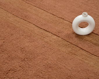 Pode incluir: Um tapete cor de terracota com listras diagonais e um vaso branco em forma de donut. O tapete tem uma superfície macia e texturizada. O vaso tem uma pequena abertura no topo.