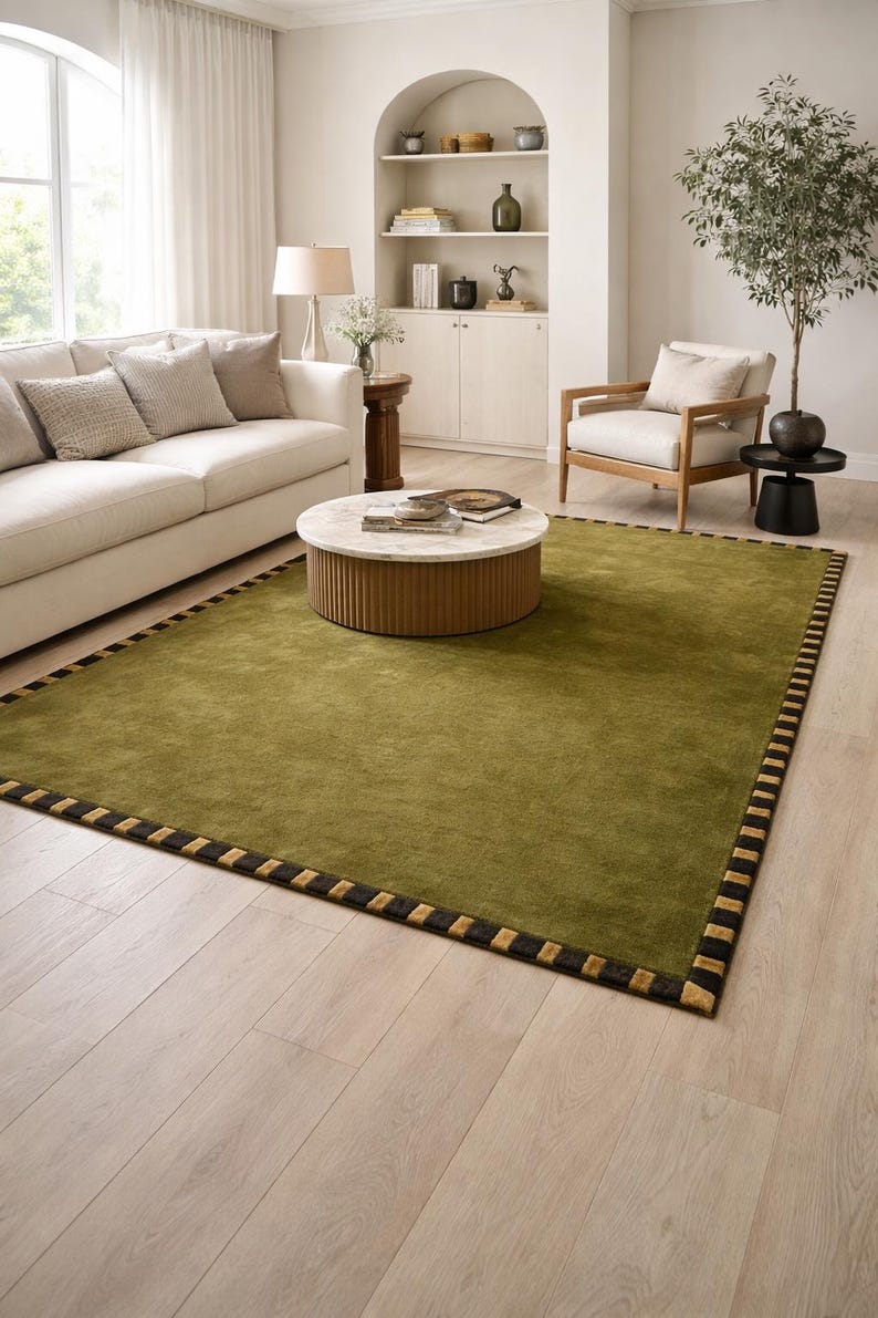 Peut inclure: Tapis rectangulaire vert olive avec une bordure noire et dor&eacute;e. Le tapis est dans un salon avec un canap&eacute; cr&egrave;me, une chaise en bois et une table basse ronde. Dimensions en cm.
