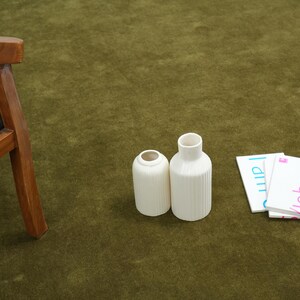 Peut inclure: Deux vases en c&eacute;ramique blanche avec des d&eacute;tails c&ocirc;tel&eacute;s et une petite pile de cartes blanches avec les noms "Johnny" et "I am" imprim&eacute;s dessus. Les articles sont sur une surface verte.