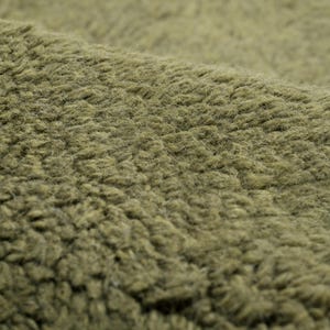 Pode incluir: Close-up de um tecido verde-oliva texturizado. O material tem um pelo denso e difuso, criando uma apar&ecirc;ncia macia e felpuda. A superf&iacute;cie do tecido &eacute; irregular, com fibras vis&iacute;veis e um ligeiro brilho. A textura geral sugere um tecido quente e confort&aacute;vel.