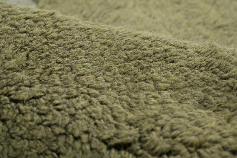 Pode incluir: Close-up de um tecido verde-oliva texturizado. O material tem um pelo denso e difuso, criando uma apar&ecirc;ncia macia e felpuda. A superf&iacute;cie do tecido &eacute; irregular, com fibras vis&iacute;veis e um ligeiro brilho. A textura geral sugere um tecido quente e confort&aacute;vel.