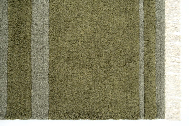 Pode incluir: Close-up de um tapete verde-oliva texturizado com duas listras cinza verticais. O tapete tem uma apar&ecirc;ncia felpuda e uma franja na borda direita. Este tapete &eacute; adequado para decora&ccedil;&atilde;o de casa.