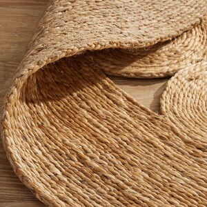 May include: A natural fiber rug made from jute, featuring a circular design with a woven texture. The rug is a light brown color and has a slightly rough surface.