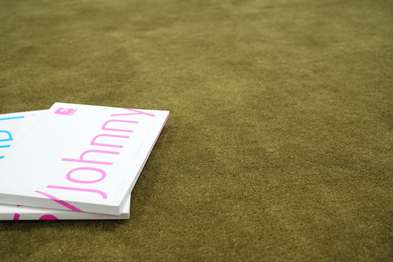 Peut inclure: Une pile de livres blancs avec le mot "Johnny" en lettres roses. Les livres sont pos&eacute;s sur une surface textur&eacute;e vert olive. Le mot "Johnny" est en gras, en police sans-serif.