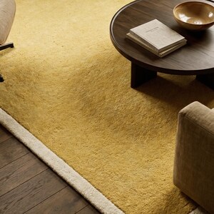 May include: A textured mustard yellow area rug with a thick cream-colored border. The rug is placed on a dark wood floor, partially covered by a round dark wood coffee table holding a stack of two books and a single light brown bowl.