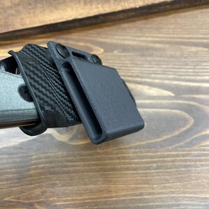 Puede incluir: Un cintur&oacute;n de cuero negro con una funda de pl&aacute;stico negro unida a &eacute;l. La funda tiene una superficie texturizada y una ranura para un cuchillo.