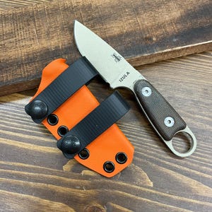 Op de afbeelding: Een klein mes met vast blad, met een tan-kleurig blad en een bruin handvat, naast een oranje schede met zwarte bandjes. Het blad heeft het woord "IZULA" en een spinnenlogo. Het mes en de schede liggen op een houten oppervlak.