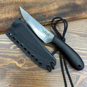 May include: A stainless steel knife with a serrated edge and a black handle, accompanied by a black leather sheath. The knife has the text "ROACH BELLY" etched on the blade. A black cord is attached to the handle and sheath.