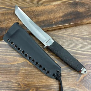 May include: A stainless steel tanto knife with a black textured handle and a black sheath. The knife blade is straight with a flat tip. The sheath has multiple eyelets and a cord. The text "MASTER TOKYO" is visible on the blade.