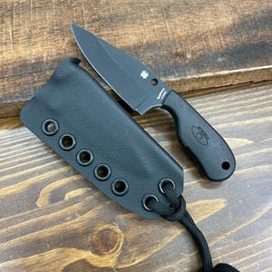 May include: A black fixed-blade knife with a matching sheath. The knife has a curved handle and a sharp blade. The sheath is made of black leather and has several eyelets. The knife and sheath are on a wooden surface.