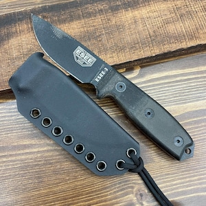 May include: A black ESEE-3 fixed blade knife with a matching black sheath. The knife has a textured handle and a partially serrated blade. The sheath has multiple eyelets and a cord for attachment. The knife is designed for outdoor use.