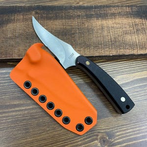 Könnte beinhalten: Ein Messer mit feststehender Klinge, einer silbernen Stahlklinge mit der Aufschrift 1520T, einem schwarzen Griff mit zwei Metallnieten und einer leuchtend orangefarbenen, strukturierten Scheide mit acht schwarzen Ösen an der Seite.