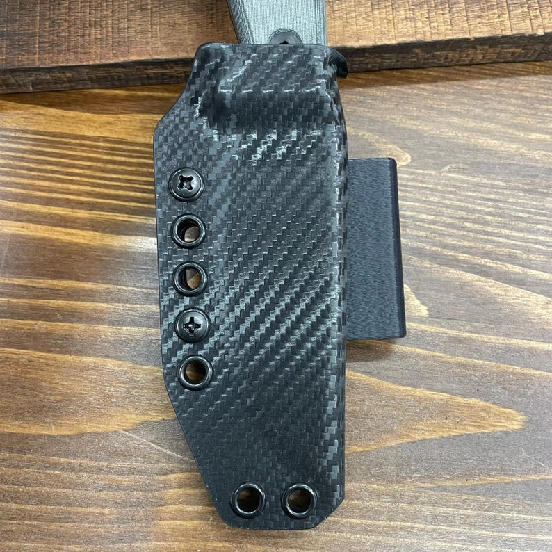 Puede incluir: Una funda para cuchillo de fibra de carbono negra con un bucle de cintur&oacute;n de cuero negro. La funda tiene varias filas de remaches y un tornillo en el lateral.