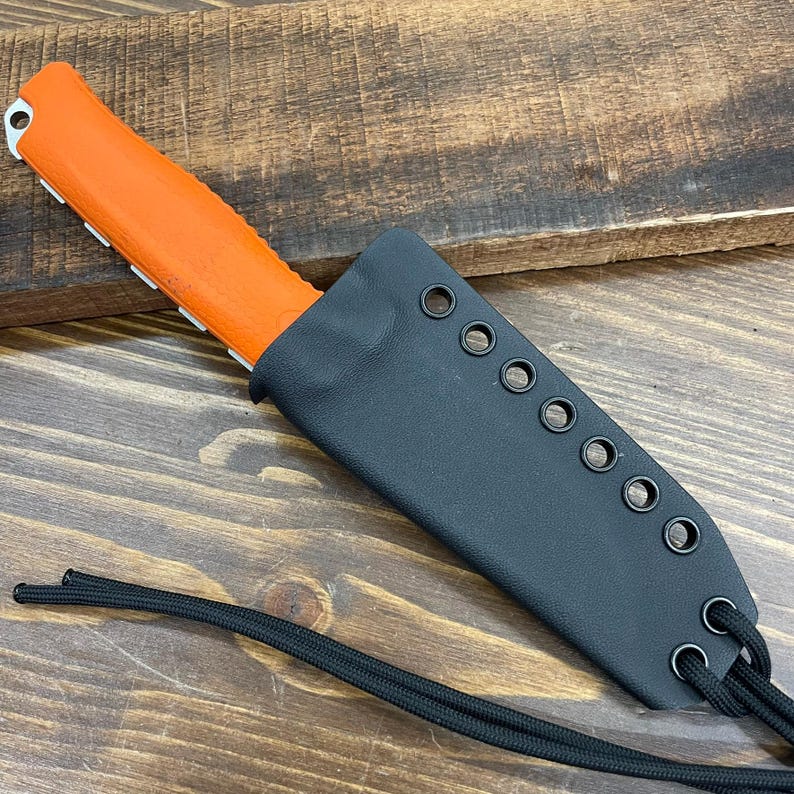 Puede incluir: Un cuchillo con mango naranja y hoja dentada est&aacute; parcialmente insertado en una funda de cuero negro. La funda tiene m&uacute;ltiples ojales a lo largo del borde y un cord&oacute;n negro. El cuchillo y la funda est&aacute;n sobre una superficie de madera.