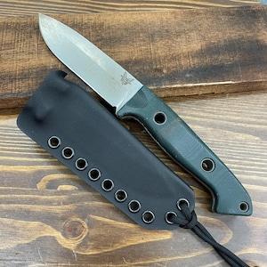 Può includere: Un coltello a lama fissa con una lama argentata e un manico verde scuro, accompagnato da un fodero in pelle nera. Il coltello presenta un logo a farfalla ed è esposto su una superficie di legno.