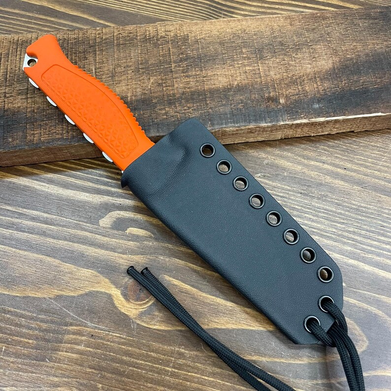 May include: An orange and black knife with a black sheath. The knife has a textured orange handle and a silver blade. The black sheath has eyelets along the edge and a black cord. The knife is on a wooden surface.