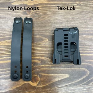 以下が含まれることがあります： 黒いナイロンループ2本と黒いTek-Lokアタッチメントデバイス。ナイロンループはテクスチャ加工された表面で、「Nylon Loops」と表示されています。Tek-Lokには一連の長方形のスロットがあり、「Tek-Lok」と表示されています。