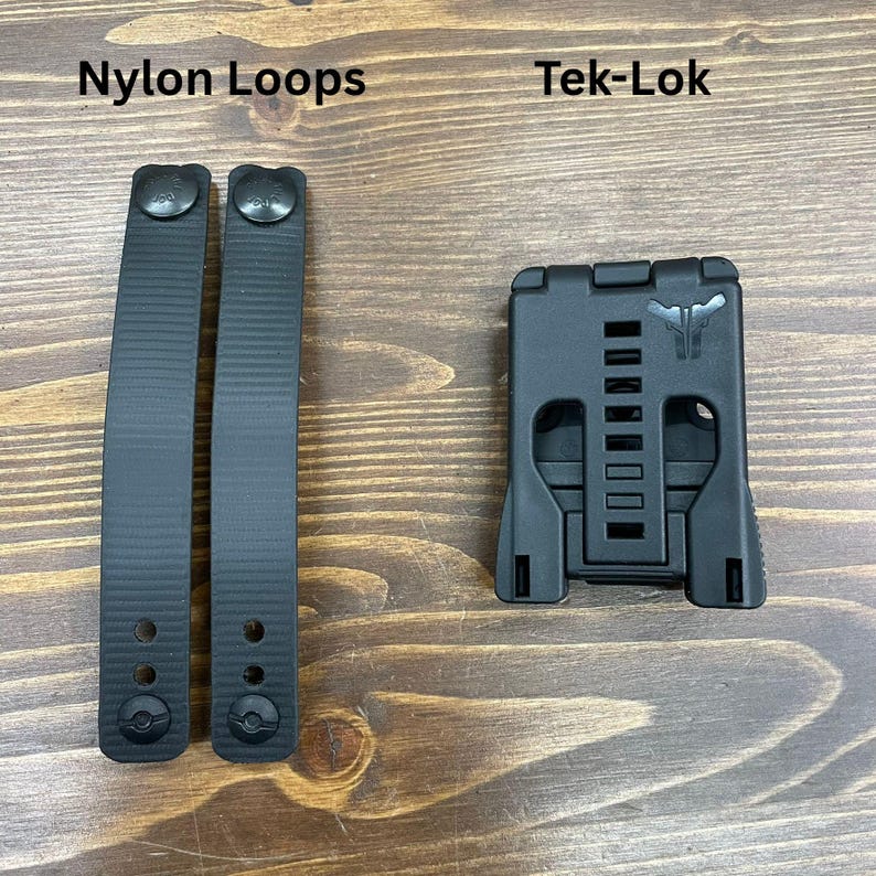 以下が含まれることがあります： 黒いナイロンループ2本と黒いTek-Lokアタッチメントデバイス。ナイロンループはテクスチャ加工された表面で、「Nylon Loops」と表示されています。Tek-Lokには一連の長方形のスロットがあり、「Tek-Lok」と表示されています。