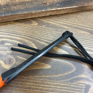 May include: Close-up of a black leather knife sheath with an orange handle. The sheath has a series of small holes along its length and is attached to a black cord. The background is a wooden surface.