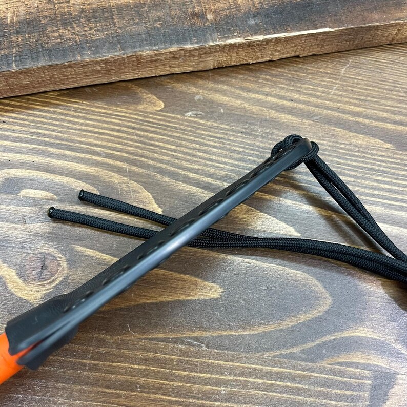 May include: Close-up of a black leather knife sheath with an orange handle. The sheath has a series of small holes along its length and is attached to a black cord. The background is a wooden surface.