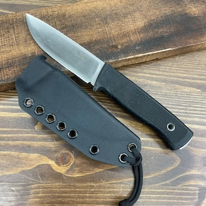 May include: A stainless steel fixed-blade knife with a black textured handle and a black sheath. The knife is lying on a wooden surface. The sheath has multiple eyelets and a cord.
