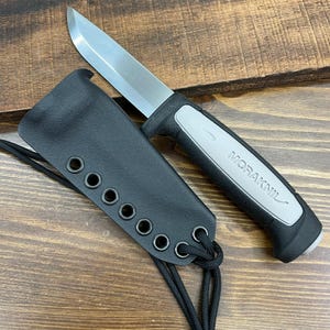 May include: A stainless steel blade knife with a black and gray handle and a black sheath. The knife is labeled "MORAKNIL" and has a lanyard attached to the sheath. The knife and sheath are on a wooden surface.