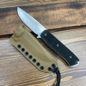 May include: A stainless steel knife with a black textured handle and a tan sheath. The knife has a silver blade and is resting on a wooden surface. The sheath has multiple eyelets and a black cord.