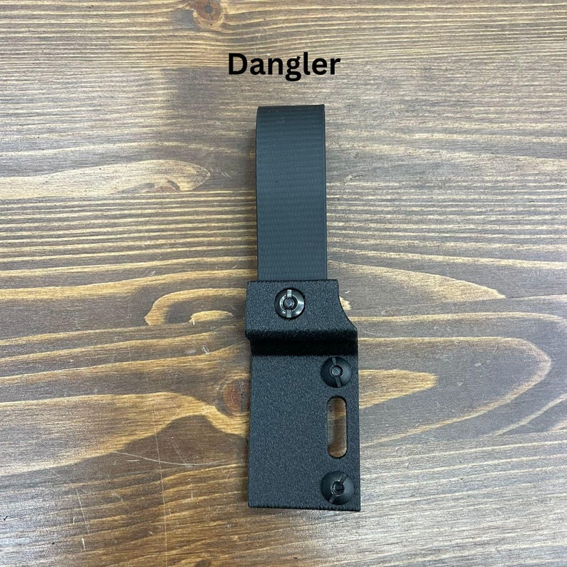以下が含まれることがあります： 黒い金属製のダンガー。黒いストラップとネジで固定されています。「Dangler」の文字が黒いテキストでオブジェクトの上に印刷されています。オブジェクトは木製の背景に配置されています。