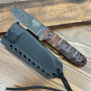 May include: A fixed-blade knife with a brown handle and a black blade, resting on a wooden surface. The knife is accompanied by a black sheath with eyelets and a black cord. The blade has the text "ESEE" and "ESEE PR4".