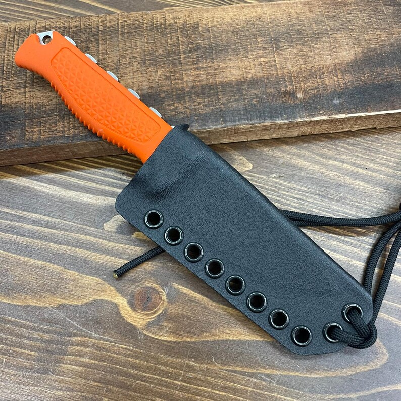 May include: An orange-handled knife with a black sheath. The knife has a textured handle and a silver blade. The black sheath has multiple eyelets along the edge and a black cord. The knife is designed for outdoor use.