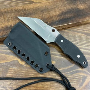 Puede incluir: Un cuchillo de acero inoxidable con mango negro y funda negra. El cuchillo tiene una forma de hoja única y está sobre una superficie de madera. La funda tiene múltiples ojales y un cordón.