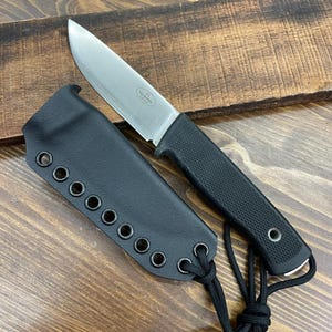 May include: A stainless steel knife with a black textured handle and a black sheath. The knife blade is silver and has a pointed tip. The sheath has multiple eyelets and a black cord. The knife is on a wooden surface.