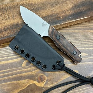 May include: A small, silver blade knife with a wooden handle and a black leather sheath. The knife has a serrated edge and a butterfly logo. The sheath has multiple eyelets and a black cord. The knife is on a wooden surface.
