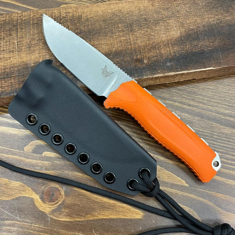 Puede incluir: Un cuchillo con mango naranja, hoja dentada y funda negra. El cuchillo tiene una hoja plateada y un mango naranja texturizado. La funda negra tiene m&uacute;ltiples ojales y un cord&oacute;n negro. El cuchillo est&aacute; sobre una superficie de madera.