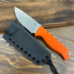 May include: A stainless steel knife with an orange handle and a black sheath. The knife blade has a serrated edge. The sheath is black with eyelets and a black cord. The knife is on a wooden surface.