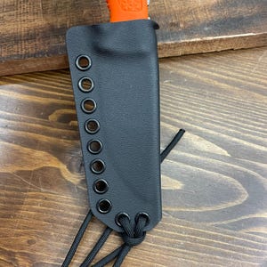May include: Black leather knife sheath with a series of metal grommets along the side for lacing. The sheath has a black cord tied at the bottom. The top of an orange knife handle is visible.