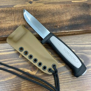 May include: A stainless steel blade knife with a black and gray handle. The knife is next to a tan sheath with black eyelets and a black cord. The knife handle has the word "MORAKNIL" printed on it.