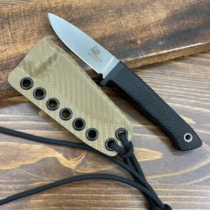 Könnte beinhalten: Ein kleines Messer mit silberner Klinge und einem schwarzen, strukturierten Griff. Dazu gehört eine beige Scheide mit Ösen und einer schwarzen Kordel. Das Messer ist mit "TITENDLETON MINI HUNTER" beschriftet.