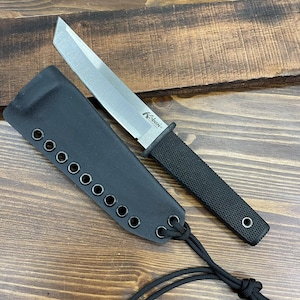 May include: A black knife with a silver blade and a black handle, resting next to its black sheath. The knife has the brand name "Kobun" printed on the blade. The sheath has multiple eyelets and a cord.