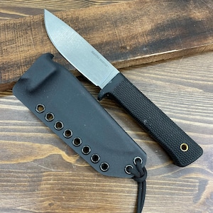 May include: A black knife with a silver blade and a textured black handle rests beside its black sheath. The blade is marked "MASTER HUNTER". The sheath has multiple eyelets and a cord. The knife and sheath are on a wooden surface.