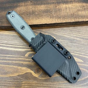 May include: A black and gray carbon fiber knife sheath with a black leather belt clip. The sheath has a textured surface and multiple holes for attaching to a belt.