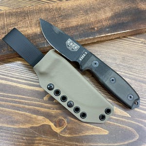 Puede incluir: Un cuchillo negro ESEE-3 con una hoja fija y un mango negro, colocado sobre una funda de color canela. El cuchillo tiene el texto "ESEE-3" y el logotipo de ESEE impreso en la hoja. La funda tiene remaches negros.