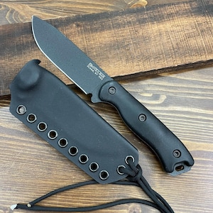 Könnte beinhalten: Ein schwarzes Messer mit passender Scheide. Das Messer hat eine schwarze Klinge und einen schwarzen Griff. Die Scheide besteht aus schwarzem Material und hat mehrere Ösen. Der Text "BK&T KA-BAR OLEAN, NY USA" ist auf der Klinge zu sehen.
