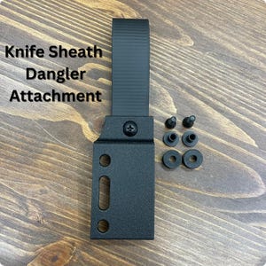 Peut inclure: Attache de support de couteau noire avec matériel de montage. L'attache a une base rectangulaire avec trois trous et une fente, et une sangle pour la fixation à une ceinture. Le texte "Knife Sheath Dangler Attachment" est imprimé sur l'image.