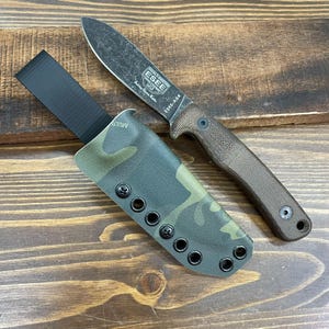 May include: A fixed-blade knife with a textured brown handle and a dark, weathered blade. The knife is accompanied by a camouflage sheath with black hardware. The text "ESEE-AGK" is visible on the blade.