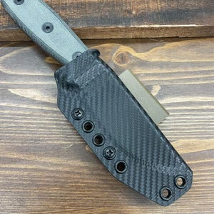 Puede incluir: Una funda de cuchillo de fibra de carbono negra con un cuchillo verde en su interior. La funda tiene varios tornillos y ojales negros.