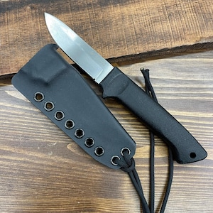 Puede incluir: Un cuchillo negro con una hoja plateada y una funda negra. El cuchillo tiene un mango texturizado y una hoja puntiaguda. La funda tiene múltiples ojales y un cordón para asegurar el cuchillo. El cuchillo y la funda están sobre una superficie de madera.