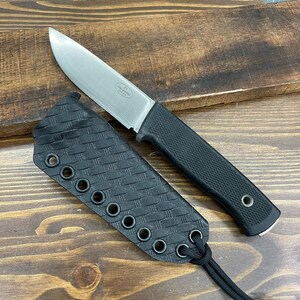 May include: A stainless steel fixed blade knife with a black textured handle and a black sheath. The knife blade is silver and the sheath has a woven pattern with multiple eyelets. The knife is on a wooden surface.