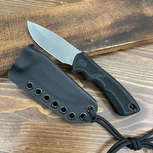 May include: A black knife with a silver blade and a black handle, resting next to a black sheath. The blade has the word "BUCK" etched on it. The knife and sheath are on a wooden surface.
